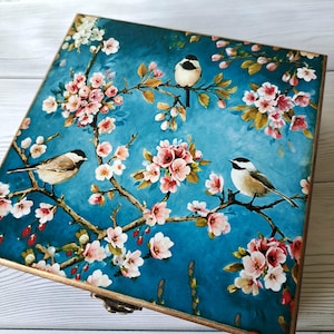 Könnte beinhalten: Quadratische Holzbox mit türkisblauem Hintergrund, verziert mit einem Blumen- und Vogelmotiv. Das Design zeigt rosa und weiße Blüten auf Ästen, mit drei kleinen Vögeln. Die Box hat eine goldfarbene Umrandung und einen Metallverschluss.