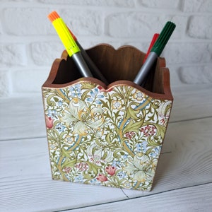 Może przedstawiać: Drewniany organizer na ołówki z kwiatowym wzorem. Organizer ma falistą górę i jest ozdobiony wzorem zielonych liści, białych kwiatów i czerwonych jagód. W organizerze znajdują się trzy markery, jeden zielony, jeden czerwony i jeden żółty.