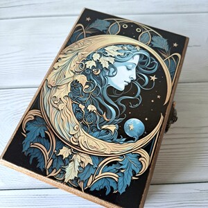 May include: A wooden box with a crescent moon design featuring a woman's face and gold accents. The background is a dark blue with stars and leaves.