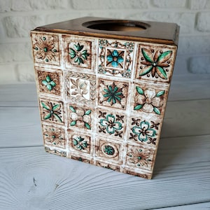 May include: Square tissue box with a dark brown wooden top and a decorative tiled design. The tiles feature floral patterns in shades of brown, white, and teal. The box is made of wood and has a square shape.
