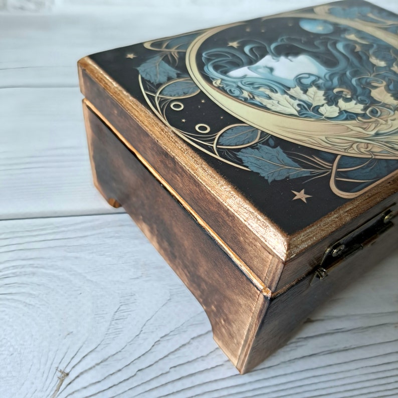 May include: A wooden box with a hinged lid. The lid is decorated with a dark blue and gold illustration of a woman's face surrounded by leaves and stars.