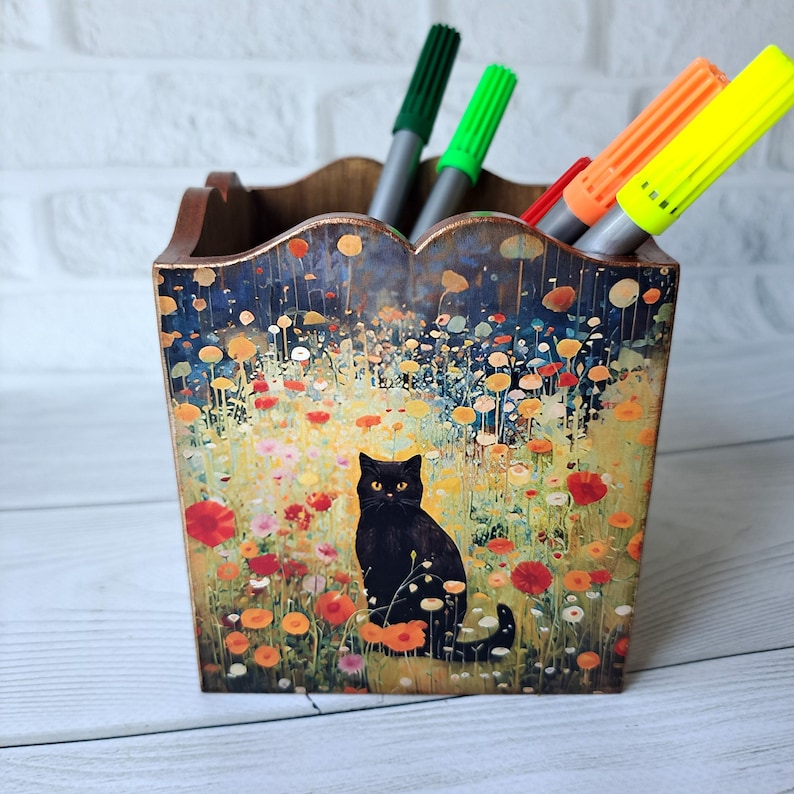 May include: A wooden pencil holder with a colorful floral design and a black cat sitting in a field of flowers. The holder has a scalloped top and is filled with colorful markers.
