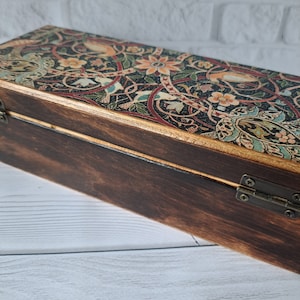 May include: A wooden box with a hinged lid. The lid is decorated with a colorful floral pattern and gold accents. The box is closed and sitting on a white wooden surface.
