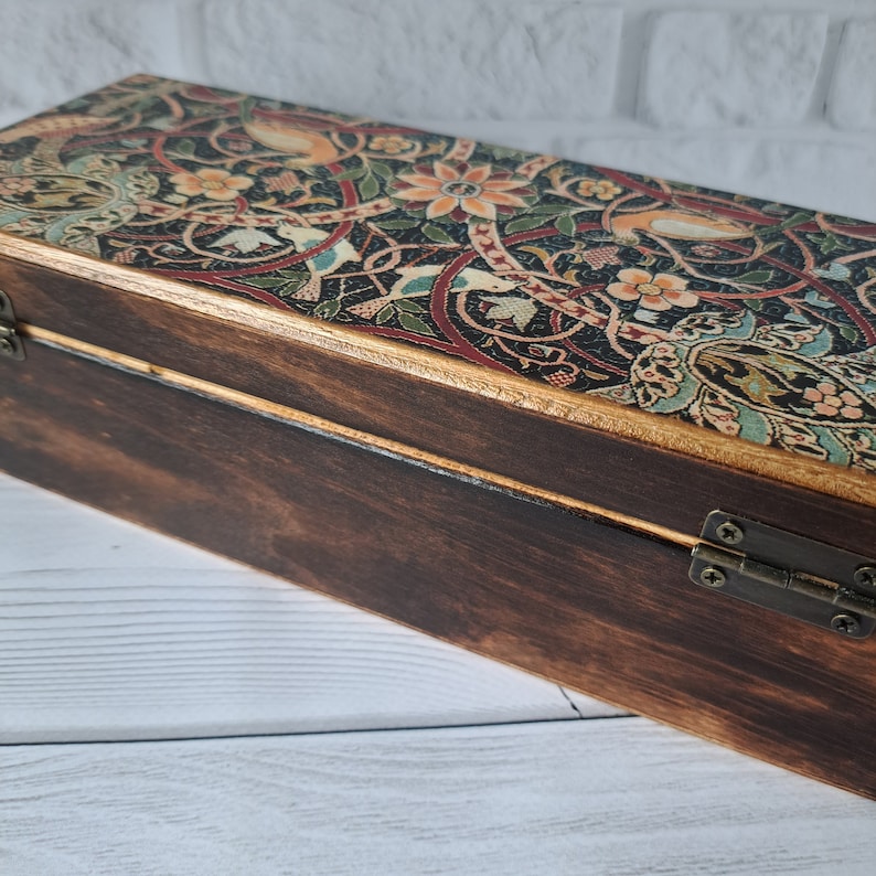 May include: A wooden box with a hinged lid. The lid is decorated with a colorful floral pattern and gold accents. The box is closed and sitting on a white wooden surface.