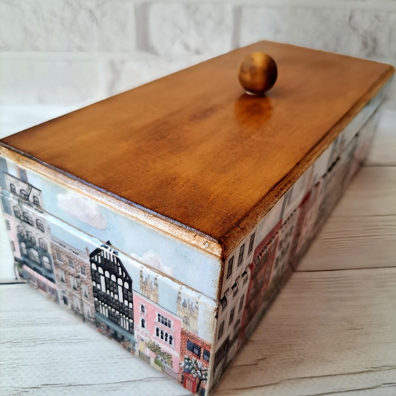 May include: A rectangular wooden box with a light brown lid and a decorative exterior. The sides of the box feature a collage of colorful buildings, creating a cityscape design. A small, round wooden knob serves as the handle on the lid.