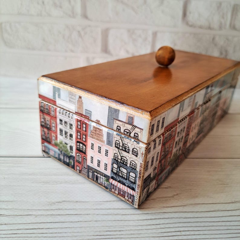 May include: A rectangular wooden box with a cityscape design. The box features a light brown, varnished lid and a decorative exterior with a repeating pattern of buildings in various colors. A small, round wooden knob serves as the handle.
