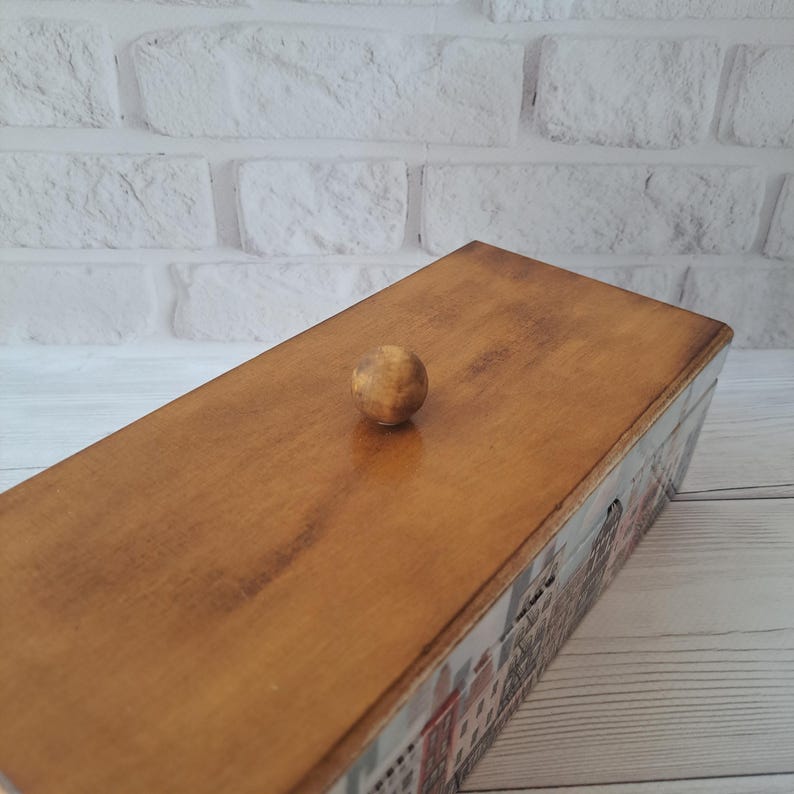 May include: A rectangular wooden box with a light brown finish and a small, round wooden knob on the lid. The sides of the box feature a colorful illustration of buildings. The box is set against a white brick wall.
