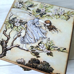 May include: A wooden box featuring a detailed illustration of a fairy in a flowing dress, with large wings, amidst flowering branches. The artwork is in soft pastel colours, with a vintage aesthetic.