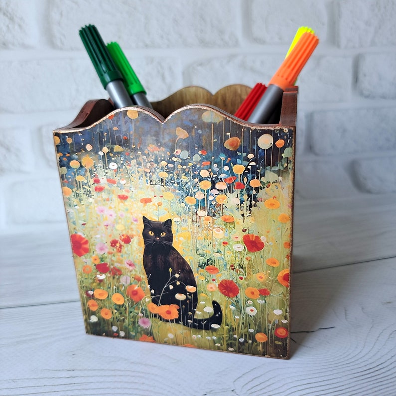 May include: A wooden pencil holder with a black cat sitting in a field of flowers. The holder has a scalloped top and is decorated with a colorful floral design.