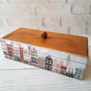 May include: A rectangular wooden box with a light brown finish and a small, round knob on top. The sides of the box are decorated with a colorful cityscape design featuring various buildings. The box is set against a white brick wall.