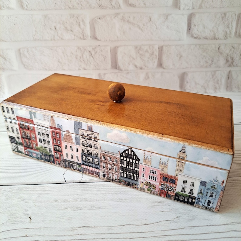May include: A rectangular wooden box with a light brown finish and a small, round knob on top. The sides of the box are decorated with a colorful cityscape design featuring various buildings. The box is set against a white brick wall.