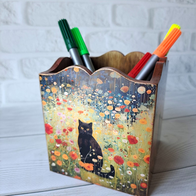 May include: A wooden pencil holder with a colorful floral design featuring a black cat. The holder has a scalloped top and is filled with three markers, one red, one green, and one neon green.