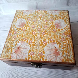 Könnte beinhalten: Holzkiste mit dekorativem Deckel mit einem Blumenmuster in Gelb-, Pfirsich- und Weißtönen. Die Kiste hat einen dunkelbraunen Sockel und einen Metallverschluss. Das florale Design umfasst stilisierte Blumen und Blätter.