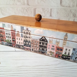 May include: A wooden box with a cityscape design. The box features a light brown wooden top and a decorative band around the sides depicting buildings, including Big Ben. A small, round wooden knob serves as the handle.