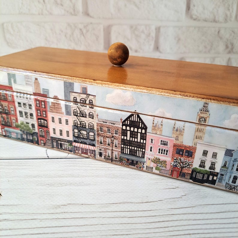 May include: A wooden box with a cityscape design. The box features a light brown wooden top and a decorative band around the sides depicting buildings, including Big Ben. A small, round wooden knob serves as the handle.