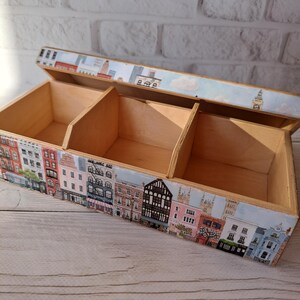 May include: Wooden storage box with three compartments, featuring a painted cityscape design. The box has a hinged lid and is decorated with colorful buildings, trees, and a blue sky. The box is made of light-colored wood.