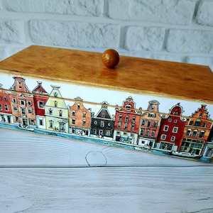 May include: Wooden box with a light brown lid and a decorative border featuring a colorful illustration of canal houses. The box has a small, round wooden knob on top.
