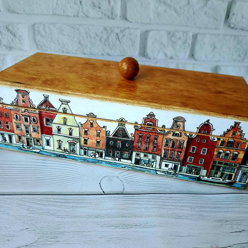 May include: Wooden box with a light brown lid and a decorative border featuring a colorful illustration of canal houses. The box has a small, round wooden knob on top.