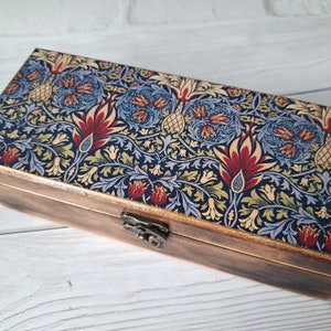 Peut inclure: Une boîte rectangulaire en bois avec un couvercle à motifs. Le couvercle présente un fond bleu foncé avec un motif floral rouge, jaune et bleu. La boîte a un fermoir et des charnières en métal.