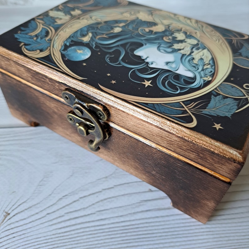 May include: A wooden box with a hinged lid and a brass latch. The lid features a colorful illustration of a woman's face with a crescent moon and stars on a dark background.