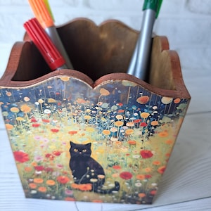 May include: A wooden pencil holder with a colorful floral design and a black cat in the center. The holder has a scalloped top and is filled with markers.