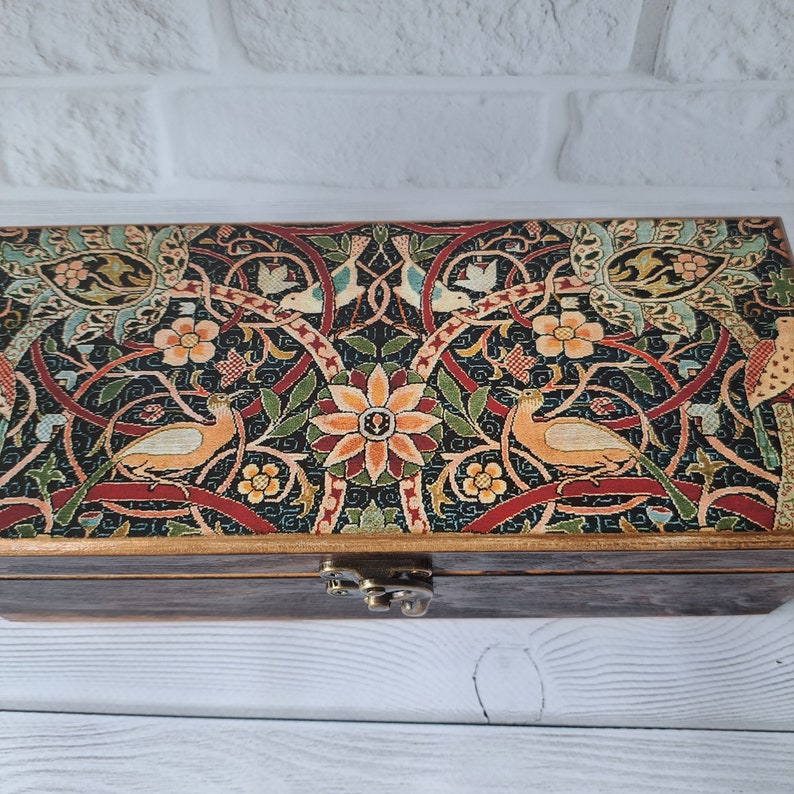 May include: A wooden box with a hinged lid. The lid is decorated with a colorful floral pattern featuring birds and intricate designs. The box has a brass latch.