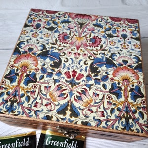 Könnte beinhalten: Eine quadratische Holzschatulle mit einem dekorativen Deckel, der ein sich wiederholendes Blumenmuster in Rosa-, Blau- und Gelbtönen auf cremefarbenem Grund zeigt. Die Schatulle hat dunkel gebeizte Seiten und einen Metallverschluss vorne.