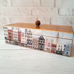 May include: A rectangular wooden box with a cityscape design. The box features a light brown wooden lid and a base adorned with a detailed illustration of buildings, including red brick structures and shops. A small, round wooden knob sits atop the lid.