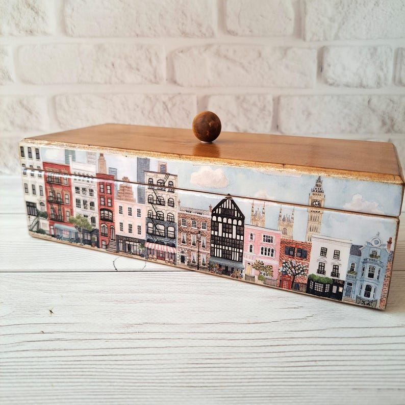 May include: A rectangular wooden box with a cityscape design. The box features a light brown wooden lid and a base adorned with a detailed illustration of buildings, including red brick structures and shops. A small, round wooden knob sits atop the lid.