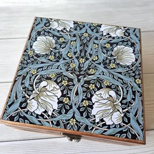 May include: A wooden box with a decorative floral design. The lid features a pattern of white flowers, light blue leaves, and yellow accents on a black background. The box has a metal clasp and a wooden frame.