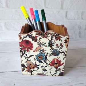 Puede incluir: Un porta lápices de madera con un diseño floral. El porta lápices está decorado con un fondo blanco y flores de colores. Hay cuatro bolígrafos en el porta lápices.