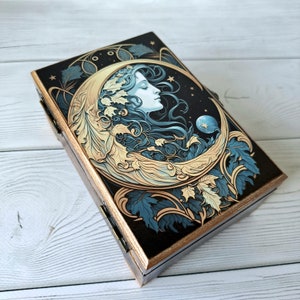 May include: A wooden box with a gold trim and a black background. The lid features a detailed illustration of a woman's face with flowing hair, surrounded by leaves and a crescent moon. The moon is gold and the leaves are blue and green.