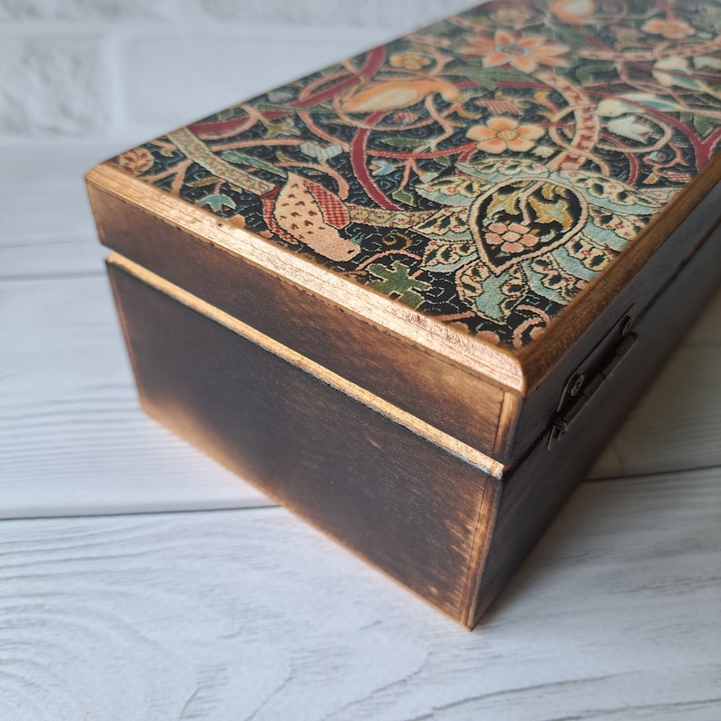 May include: A wooden box with a hinged lid. The lid is decorated with a colorful floral pattern. The box is stained a dark brown color.