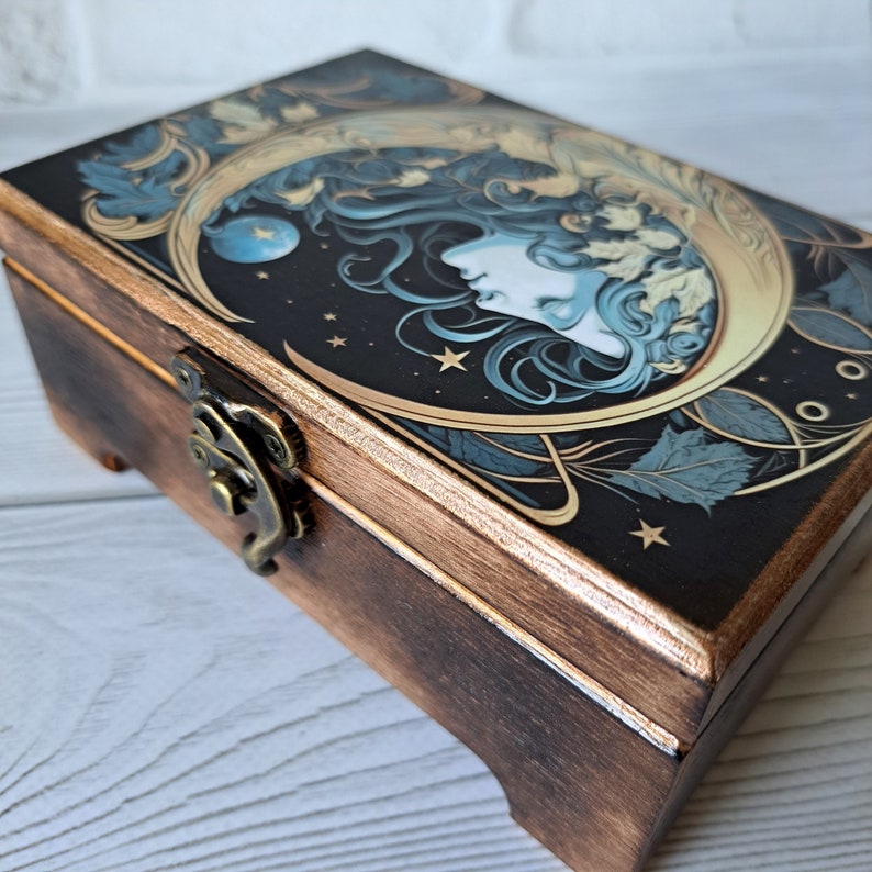 May include: A wooden box with a hinged lid and a brass latch. The lid features a colorful illustration of a woman's face with flowing hair, a crescent moon, and stars on a dark background.