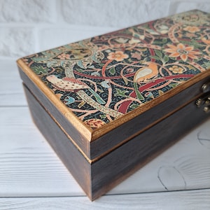 May include: A wooden box with a hinged lid. The lid is decorated with a colorful floral pattern featuring birds and intricate designs. The box has a dark brown finish and gold accents.