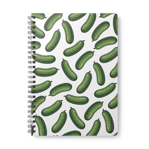 May include: A spiral-bound notebook featuring a repeating pattern of green cucumbers on a white background. The cucumbers are illustrated in a cartoon style with small dark green dots and short stems, scattered across the cover in various orientations.