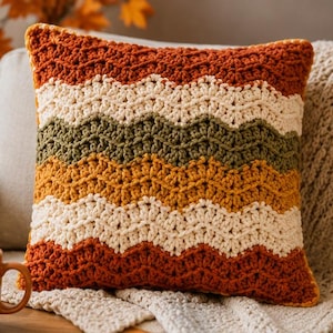 Peut inclure: Un coussin carré en crochet présentant un motif chevron horizontal avec des rayures alternées orange brûlé, crème, vert olive et jaune moutarde. Le fil texturé crée un design ondulé en zigzag sur toute la surface du coussin.