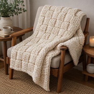 Può includere: Una coperta in maglia grossa color crema biancastro caratterizzata da un evidente motivo a intreccio. Il filato spesso crea un aspetto strutturato a griglia sull'intera superficie della coperta, che è drappeggiata su una sedia in legno.