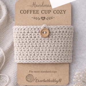 Könnte beinhalten: Handgefertigte Kaffeebecher-Hülle aus hellbeigem Häkeldesign, mit einem Holzknopf befestigt. Das Produkt ist an einem braunen Etikett mit dem Text "COFFEE CUP COZY" und "Fits most standard cups" befestigt.