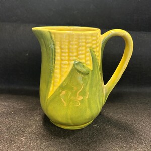 May include: A ceramic jug with a corn-on-the-cob design. The jug is yellow with green accents, featuring a handle and a spout. The design includes a detailed depiction of corn kernels and leaves.