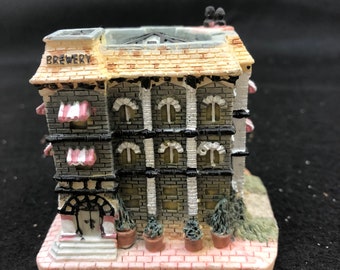 Op de afbeelding: Een miniatuurmodel van een bakstenen gebouw met een bord waarop 'Brouwerij' staat. Het gebouw heeft een grijze bakstenen gevel met witte versieringen en roze luifels. Er staan potplanten voor het gebouw.