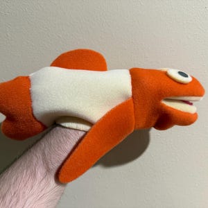 May include: Orange and white felt hand puppet shaped like a fish with a large mouth and one eye.