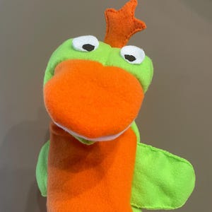 May include: Green and orange felt hand puppet with a crown on its head and white eyes. The puppet has a large orange mouth and a green wing on one side.