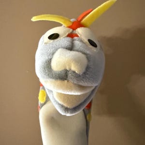 May include: A plush hand puppet featuring a cartoonish creature with a gray head, yellow antennae, and black eyes. The puppet has a light beige snout and a light beige body. The puppet's design includes red and yellow accents.