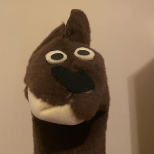 May include: A brown puppet with a black nose and white eyes. The puppet is made of soft fabric and has a long neck.