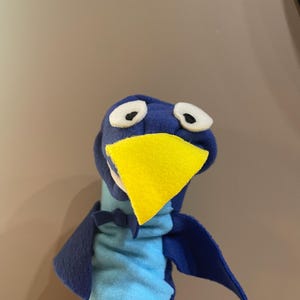 May include: Blue and light blue puppet with a yellow beak and white eyes. The puppet has a blue cape and is designed to be worn on a hand.