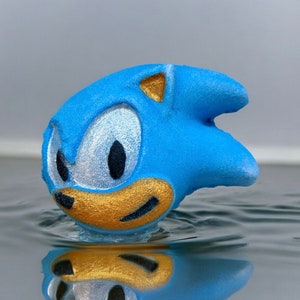 May include: A blue bath bomb shaped like Sonic the Hedgehog, a video game character. The bath bomb has gold accents and a white and blue swirl pattern.