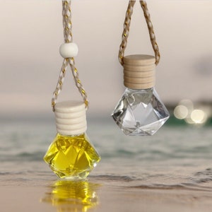 May include: Two diamond-shaped glass car air fresheners with wooden tops and string hangers. The air fresheners are filled with clear and yellow liquid.