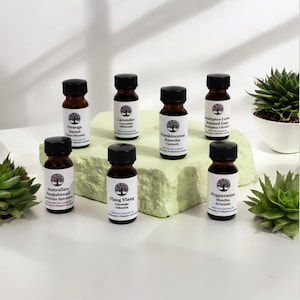 May include: Eight small brown glass bottles with labels listing different essential oils, including Orange Sweet, Lavender, Frankincense, Eucalyptus Lemon Scented Gum, Australian Sandalwood, Ylang Ylang, and Peppermint. The bottles are arranged on a green surface with two small succulent plants.