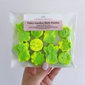 May include: A clear plastic bag containing green and yellow bath fizzies shaped like fairies, flowers, and other whimsical designs. The bag has a label that reads "Fairy Garden Bath Fizzies" and "Fragrance: Monkey Farts".